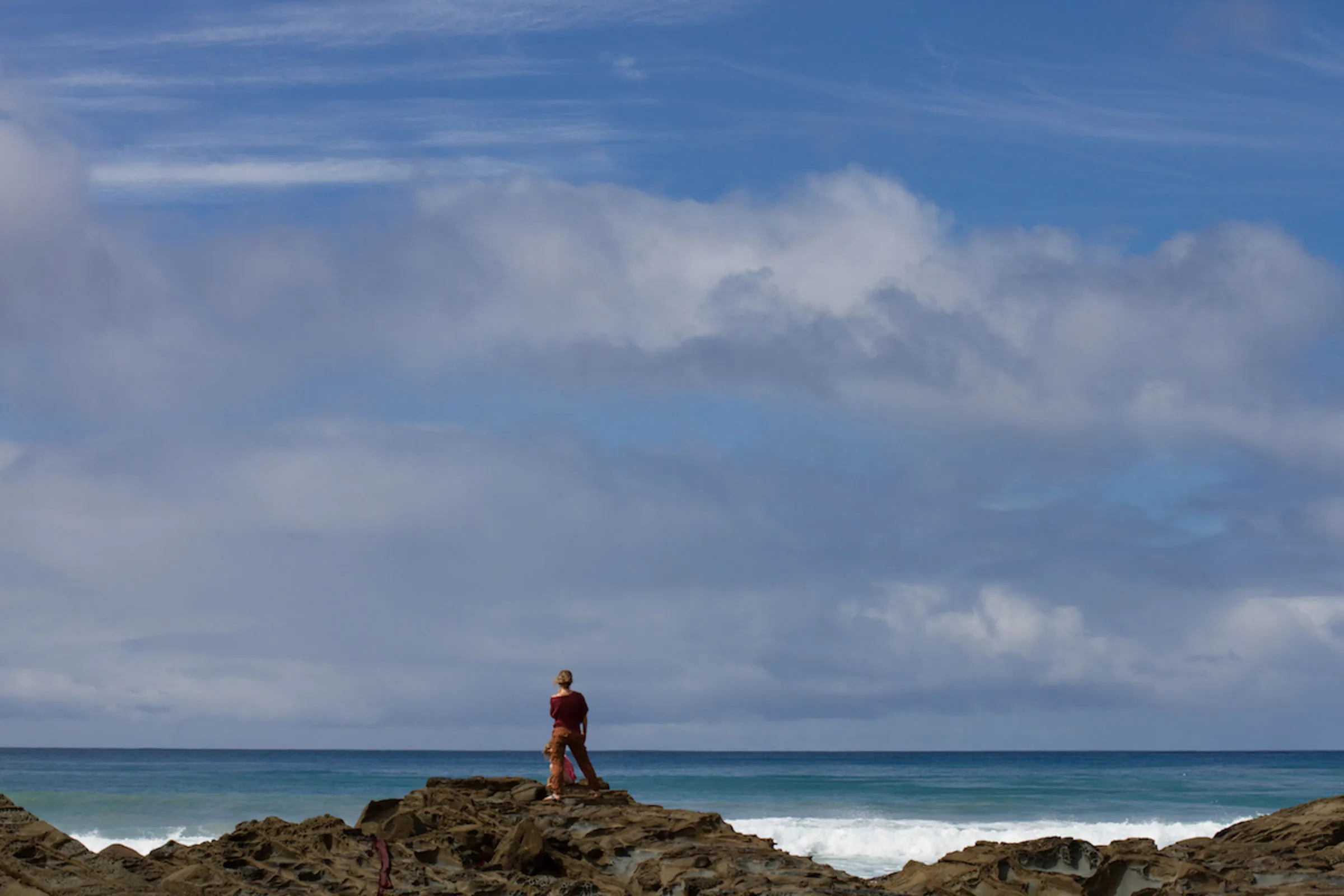 A lone figure standing on rocks above the ocean beneath a wide, cloud-streaked sky.